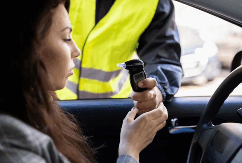 Image is of a female driver behind her driving wheel being handed a breathalyzer by a police officer outside of her car window, concept of common DUI defenses