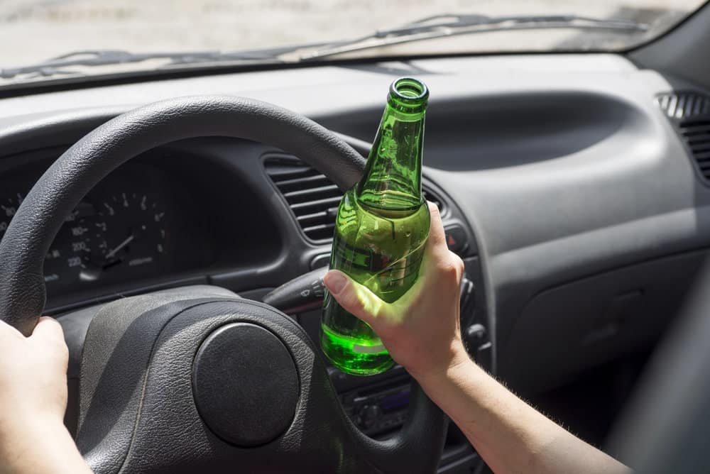 Image is of a driver holding an alcohol bottle while driving, showing risks faced by DUI and professional program students when poor decisions threaten academic and career outcomes.