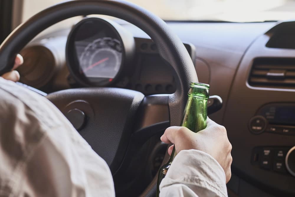 Image is of a driver holding an alcohol bottle while behind the wheel, illustrating risky behavior that can lead to DUI charges and serious legal consequences for students.