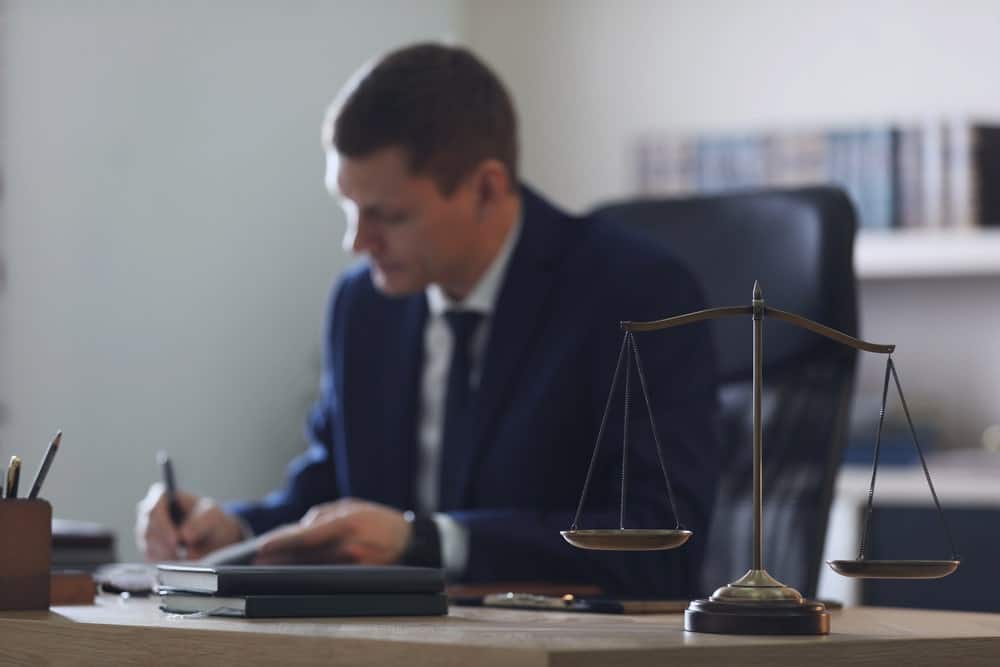 Image is of a lawyer writing notes at a desk with scales of justice in front, concept of stand your ground case legal preparation
