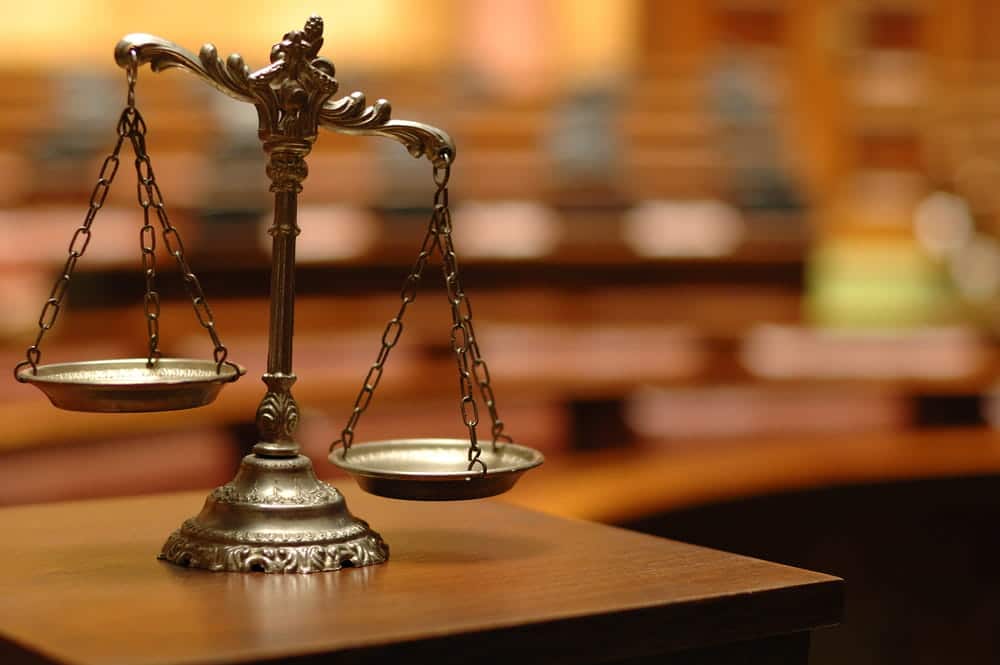Image is of silver scales of justice sitting on a wooden table in a blurred courtroom setting, representing the legal requirements for court-ordered expungement.